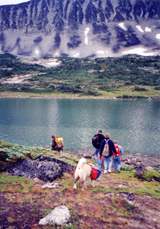 Hikers carry packs near an alpine lake.